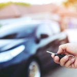Business man hand holding car keys front with car on background. parking in front of the house.