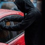 A car thief attempts to break into the window of a red car