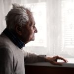 Elderly man standing indoors at home, looking out the window.
