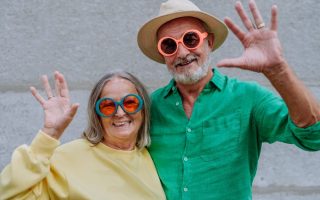 A couple of happy seniors in colourful clothes waving at camera.