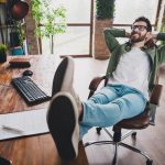 A remote worker at home, smiling and kicking up his feet on his desk